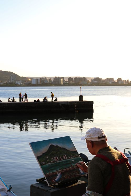 Free Artist painting a landscape by the harbor during sunset with city skyline in the background. Stock Photo