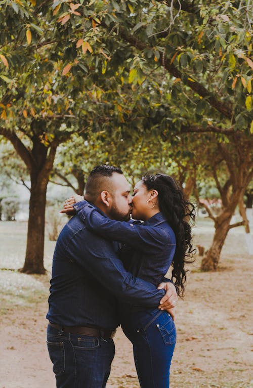 Side view of young ethnic romantic couple in similar outfits embracing and kissing each other during date in autumn park