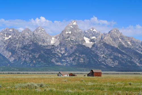Free Scenic view of rustic barns against the majestic Grand Teton mountain range under a clear blue sky. Stock Photo