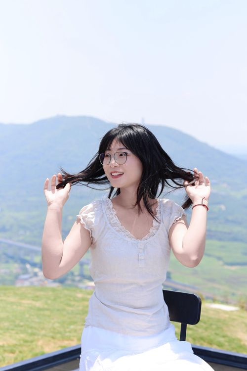 Free Smiling woman with glasses sitting outdoors, playing with hair against a scenic mountain backdrop. Stock Photo