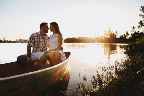 Young bearded man and woman in summer dress cuddling while sitting in traditional wooden boat on lake shore and looking at each other