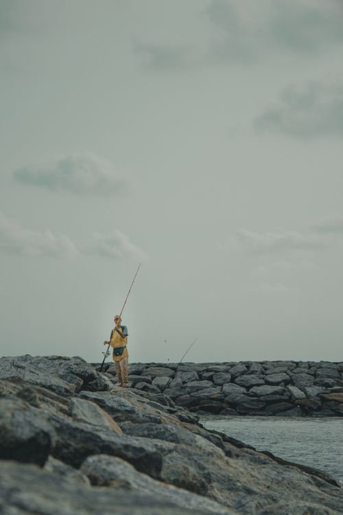 Free A person stands alone fishing on the rocky shores of Wennappuwa, Sri Lanka under a cloudy sky. Stock Photo