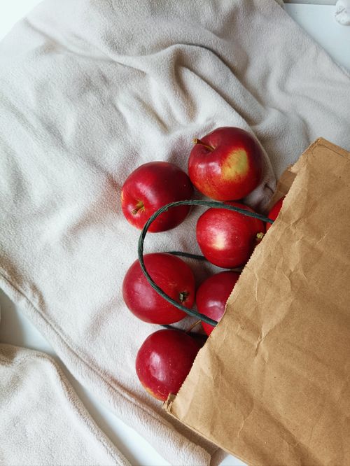 Free Aesthetic still life of fresh red apples spilling from a paper bag onto a soft beige blanket, offering a cozy and inviting atmosphere. Stock Photo