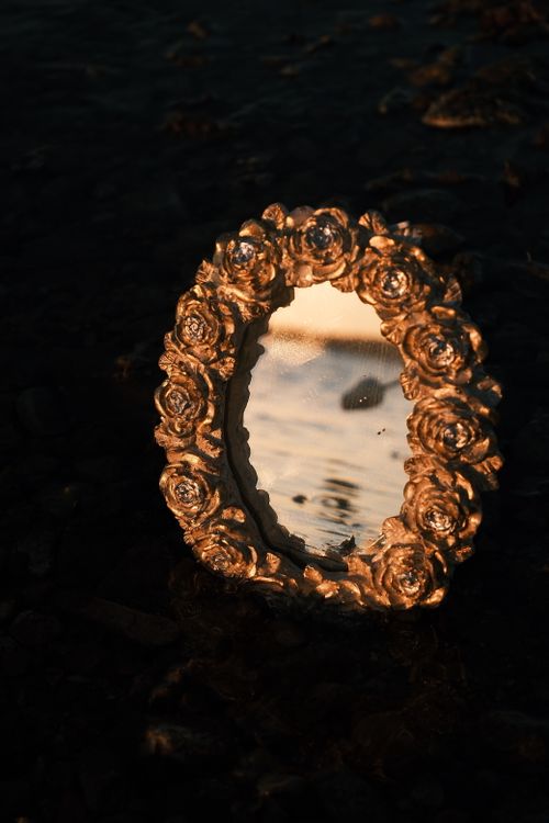 Free A decorative mirror with floral frame reflects a serene sunset over water. Stock Photo