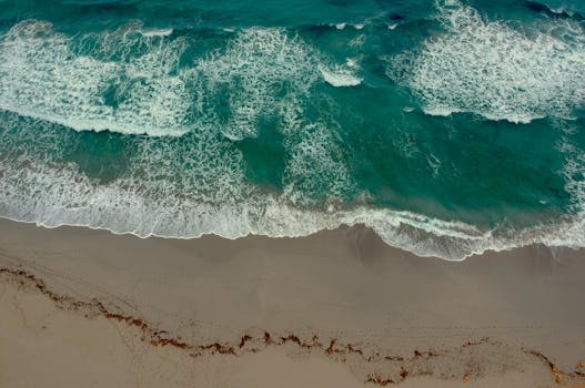 Stunning aerial capture of turquoise ocean waves crashing onto a sandy shore with footprints.
