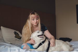 Pensive young woman with long blond hair in casual wear petting husky while resting on bed in bright room decorated in pastel colors