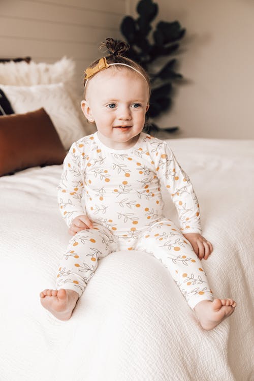 Free Full body of adorable happy little girl with bow tapped on head in cozy pajama with pattern looking away with warm smile while relaxing on bed Stock Photo