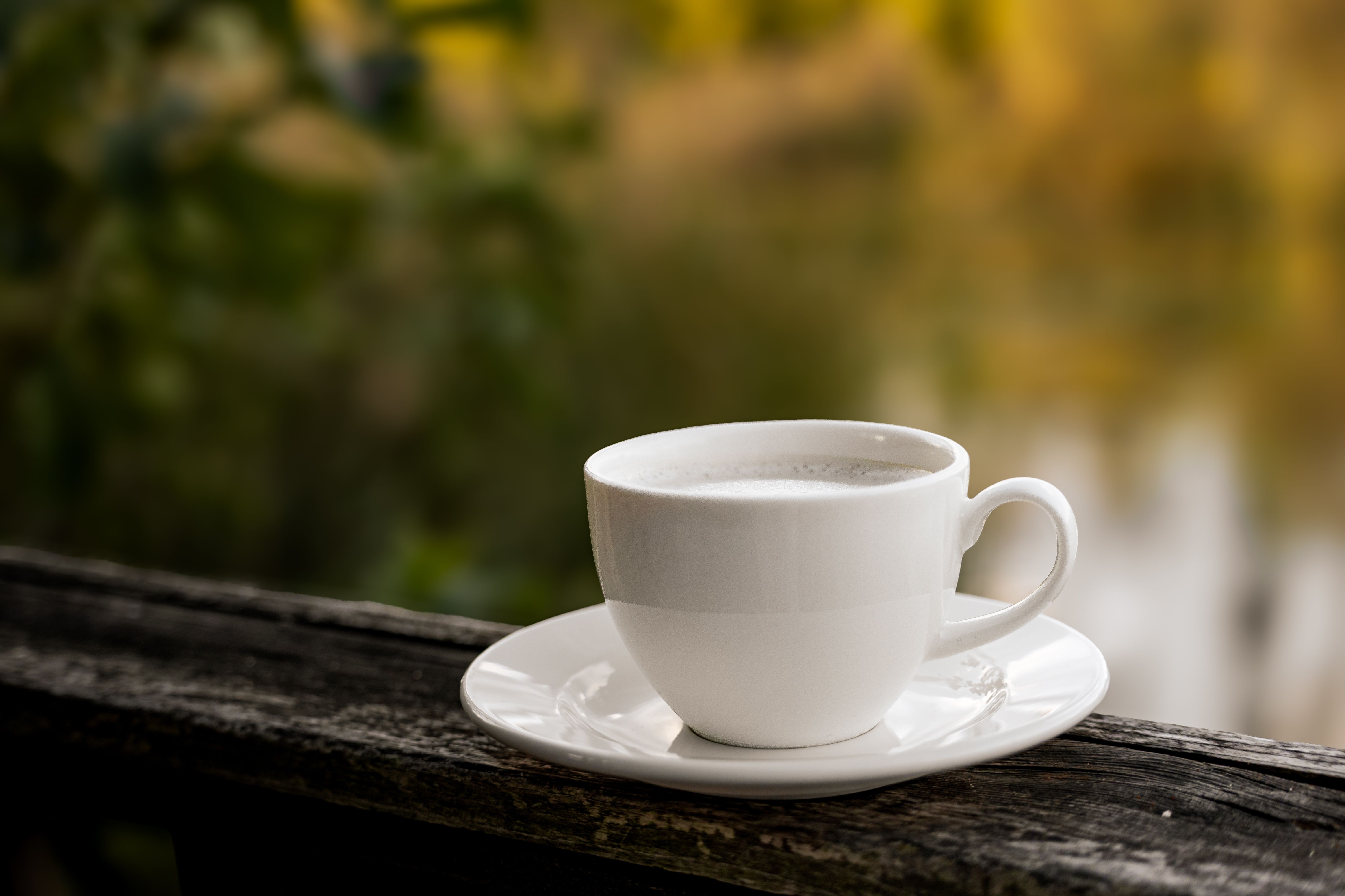 Warm cup of coffee on a rustic wooden railing with blurred autumn background.