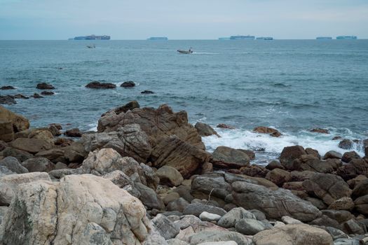 A serene view of a rocky coastline with boats and cargo ships on the horizon.