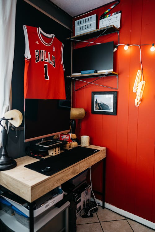 Interior of bright bedroom of adolescent with wooden table and computer decorated with garlands and black shirt attached to red wall