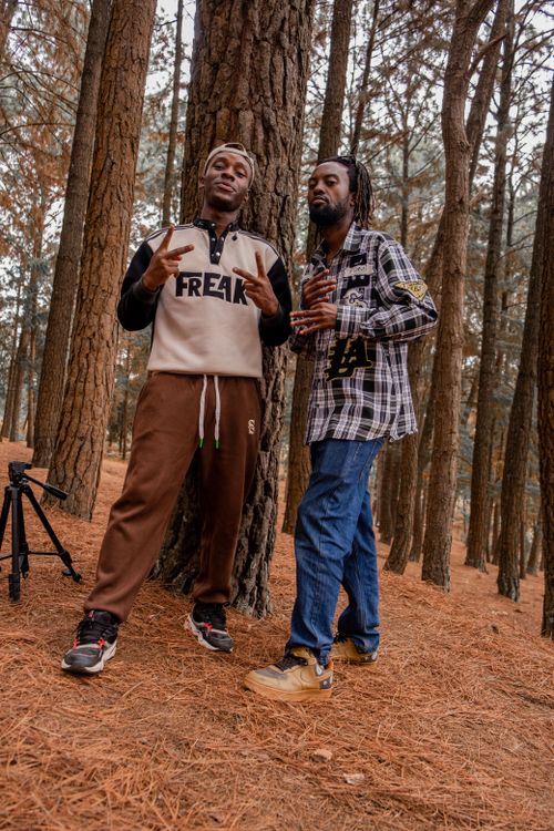 Free Two friends pose in a pine forest, showcasing their style with a camera tripod nearby. Stock Photo