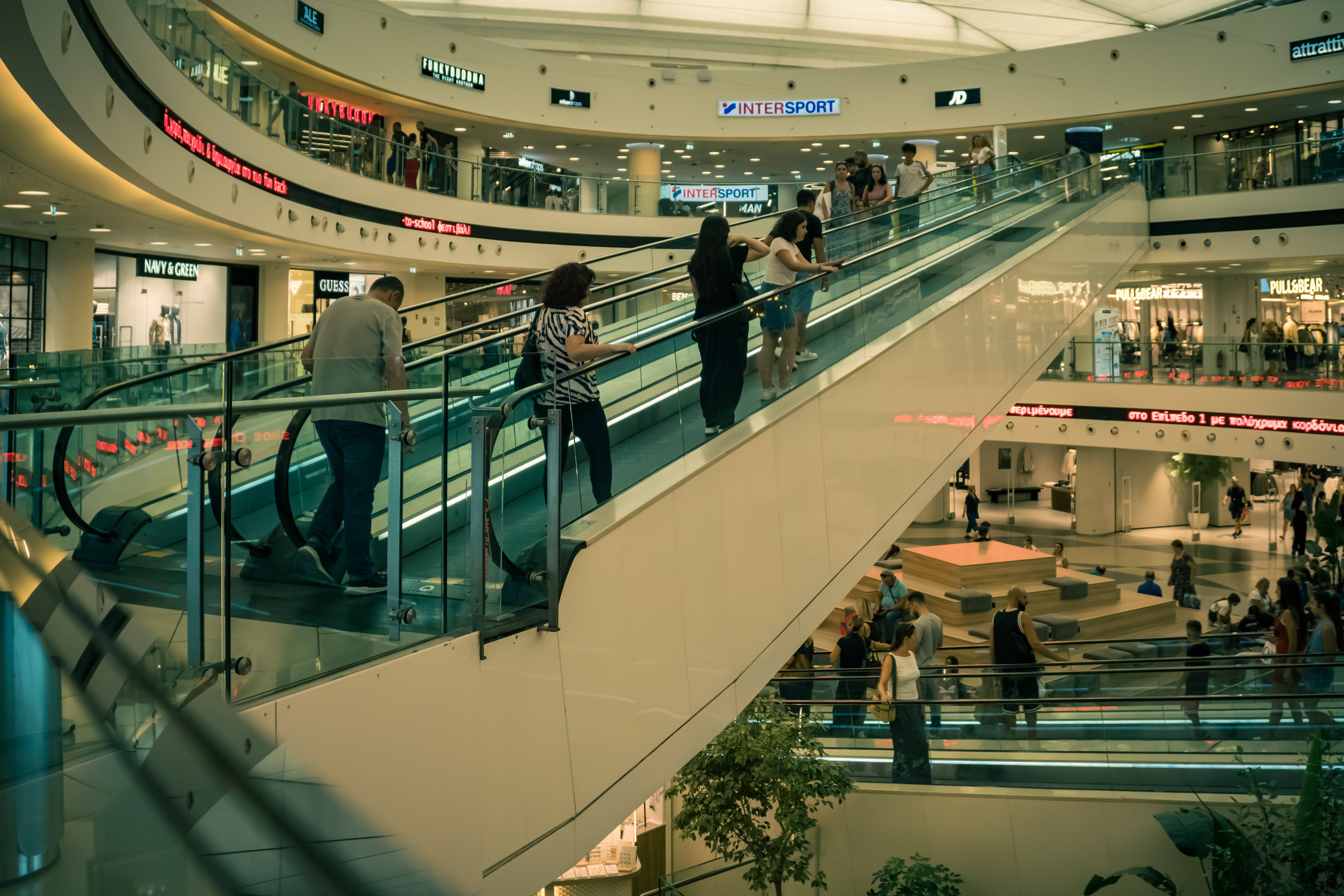 People on an escalator in a bustling contemporary shopping mall with various stores.