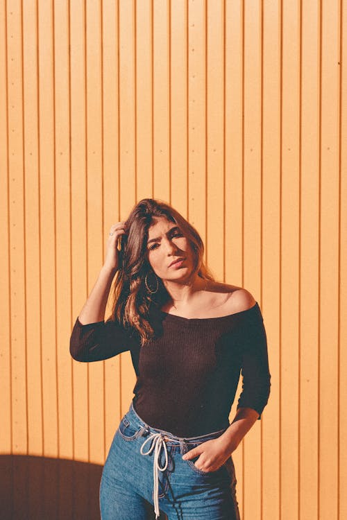 Woman in Black Scoop Neck Long Sleeve Shirt and Blue Denim Jeans Leaning on Yellow Wall