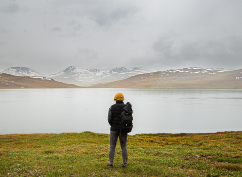 Traveler with backpack gazes at serene alpine lake and snow-capped mountains on a cloudy day.
