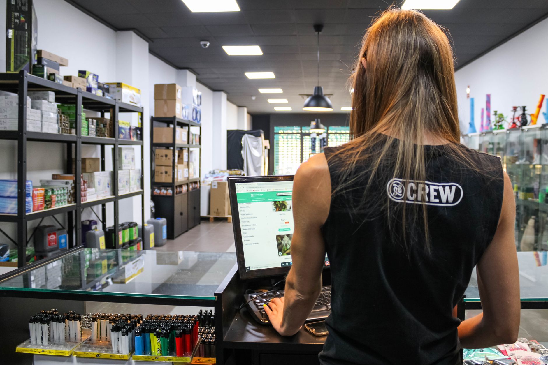 A retail worker interacts with a computer at a store counter filled with various goods.