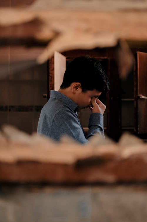 Side view of tired depressed young man with dark hair in casual clothing rubbing eyes in dark room