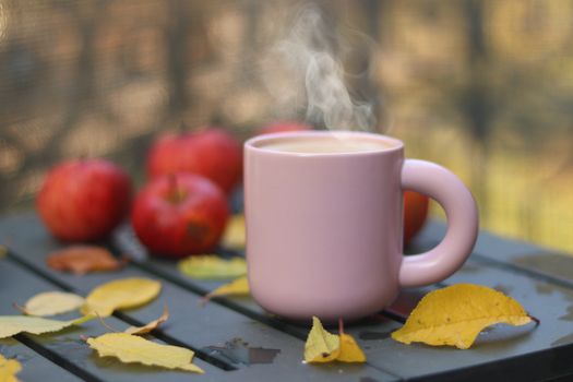 Steaming hot drink in a pink mug on a table surrounded by red apples and autumn leaves.