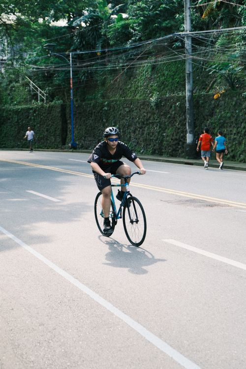 Free A cyclist riding on a sunny urban street, surrounded by greenery, capturing a vibrant city life moment. Stock Photo