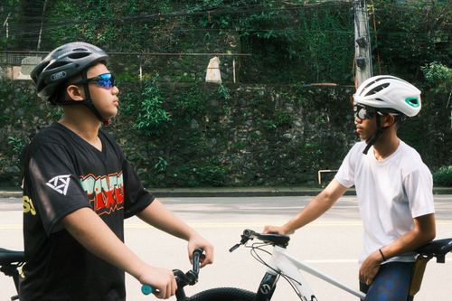 Free Two teenagers in helmets take a break on their bicycles along a leafy road, suggesting outdoor activity. Stock Photo