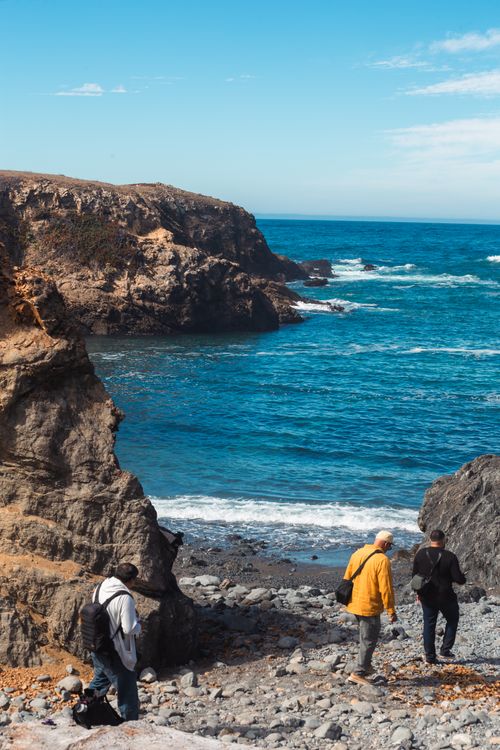 Free Three hikers explore rocky cliffs along a vibrant blue coastline under a clear sky. Stock Photo