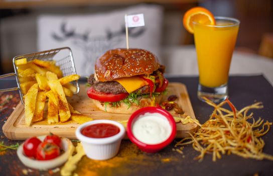 Cheeseburger with fries, sauces, and orange juice on a wooden board.