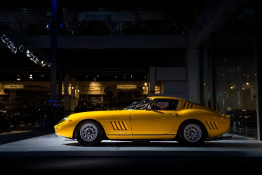 Profile view of a classic yellow sports car under dramatic showroom lighting, showcasing elegant design.