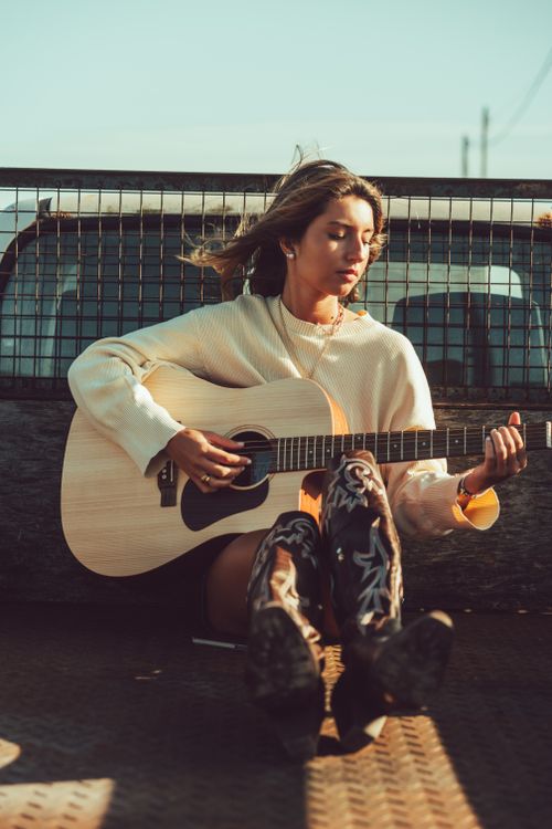 Free A stylish young woman playing acoustic guitar while seated outdoors. Perfect for lifestyle and music themes. Stock Photo