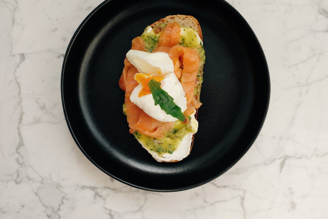 Free Top view of black ceramic plate with tasty toast with salmon and poached egg served on white marble table Stock Photo