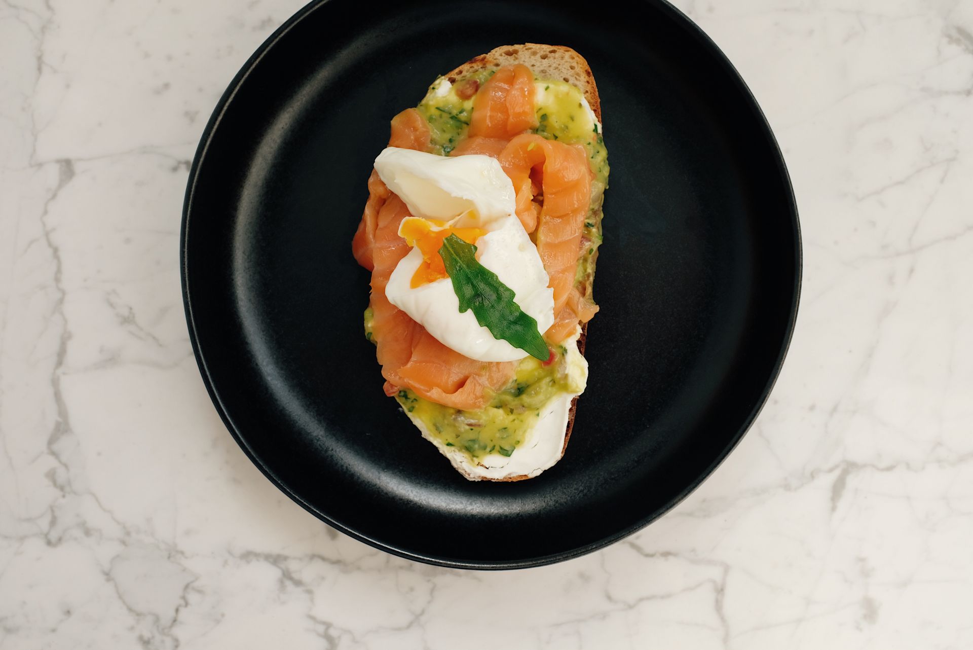 Top view of black ceramic plate with tasty toast with salmon and poached egg served on white marble table