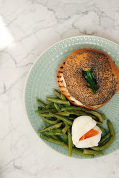 Top view of ceramic plates with toasted bagel served with pile of boiled asparagus and poached egg white marble table in light modern kitchen