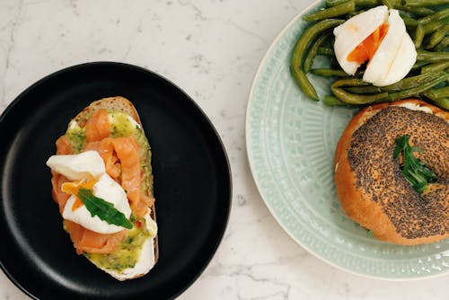 Ceramic plates with tasty burger with asparagus and poached egg and toast with fish served on white marble table in modern kitchen