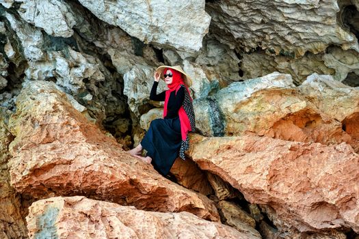 Elegant woman in a red scarf and hat sitting on rocky cliffs in Jawa Barat, Indonesia.