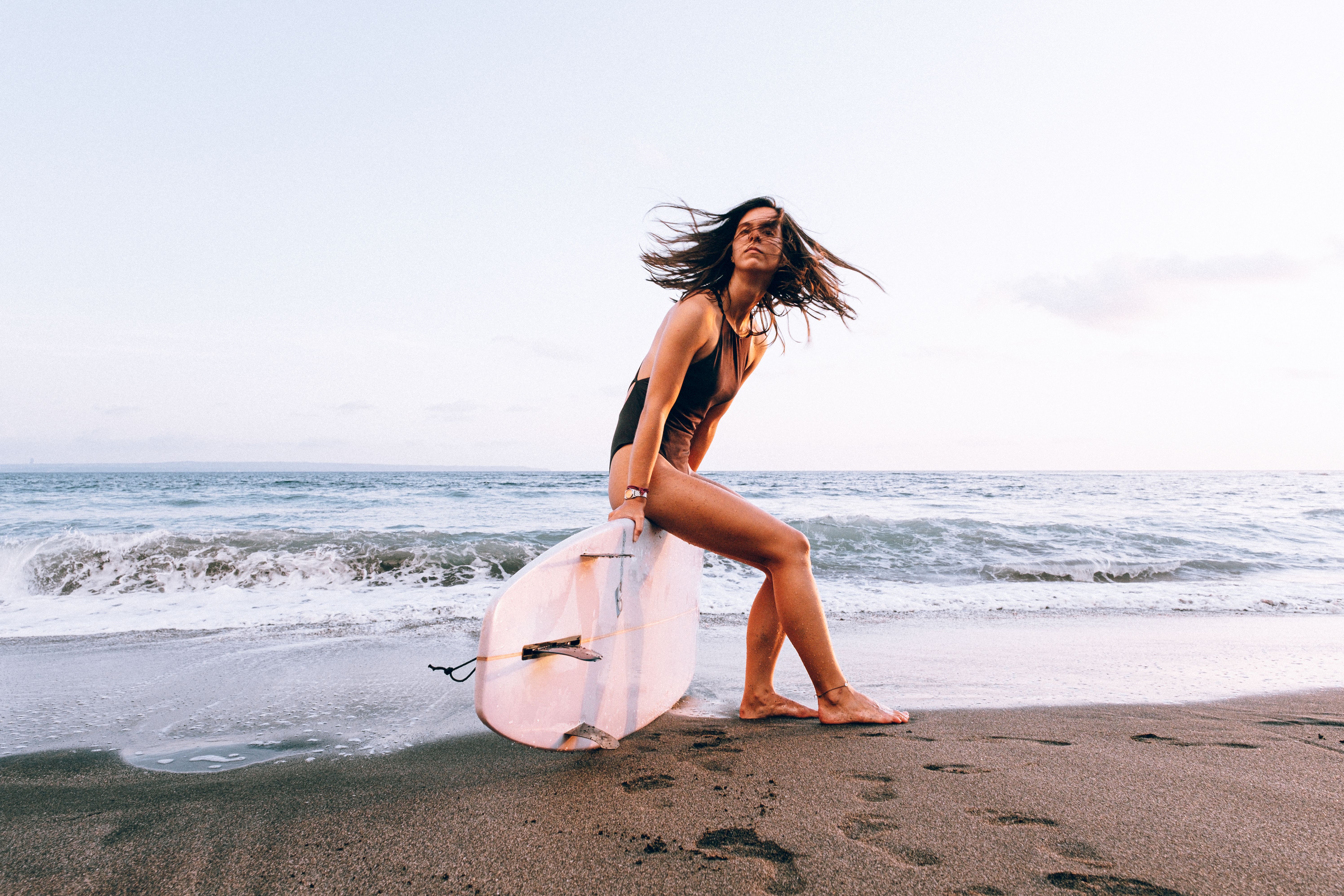 Energetic young woman posing with a surfboard on a sandy beach during sunset, ready for surfing.