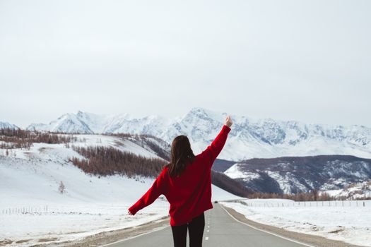 一位穿着红色毛衣的妇女站在路上，举起双臂，朝着白雪覆盖的山脉走去