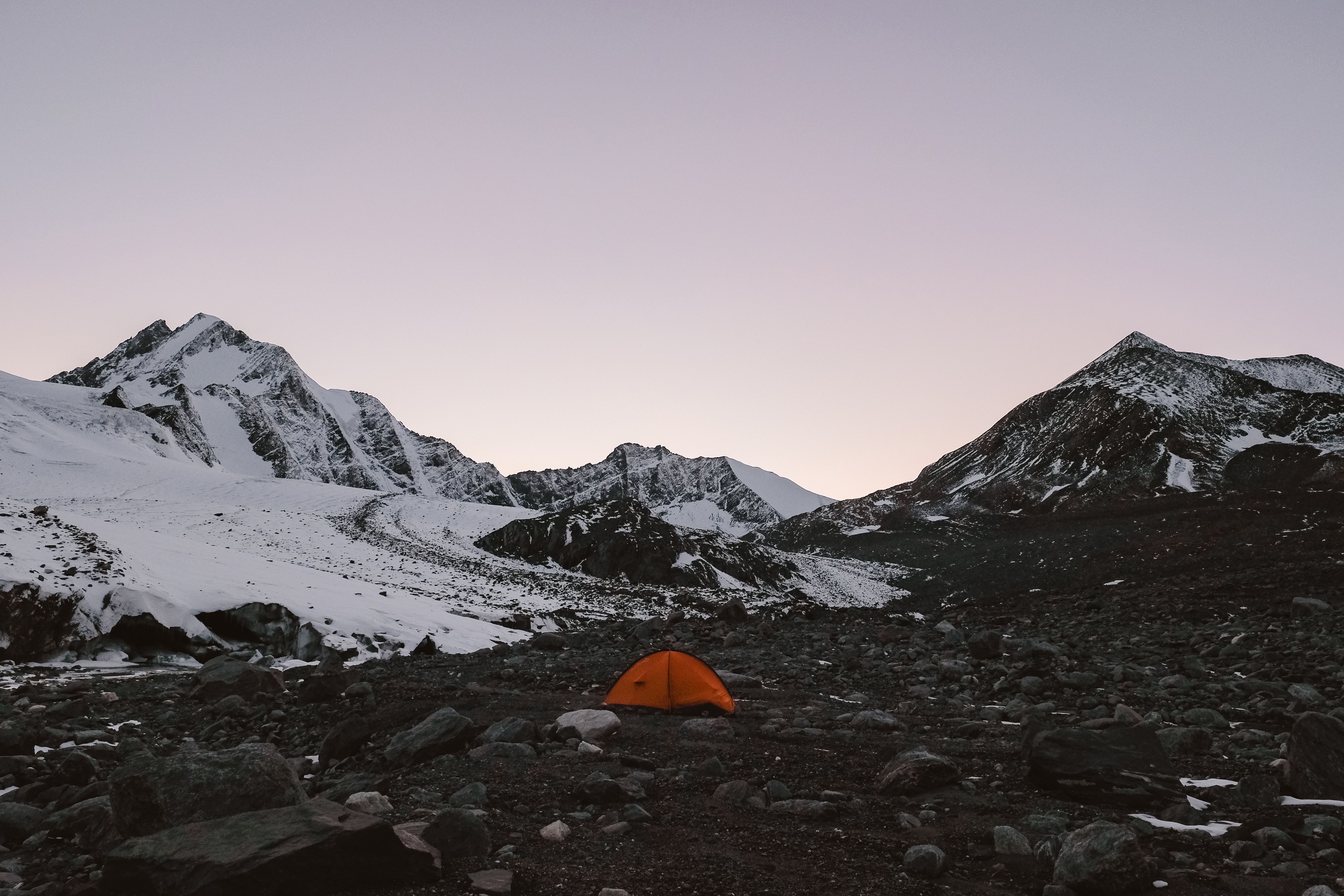 宁静的山地景观中，日落时分白雪皑皑的山峰与橙色帐篷相映，非常适合冒险主题