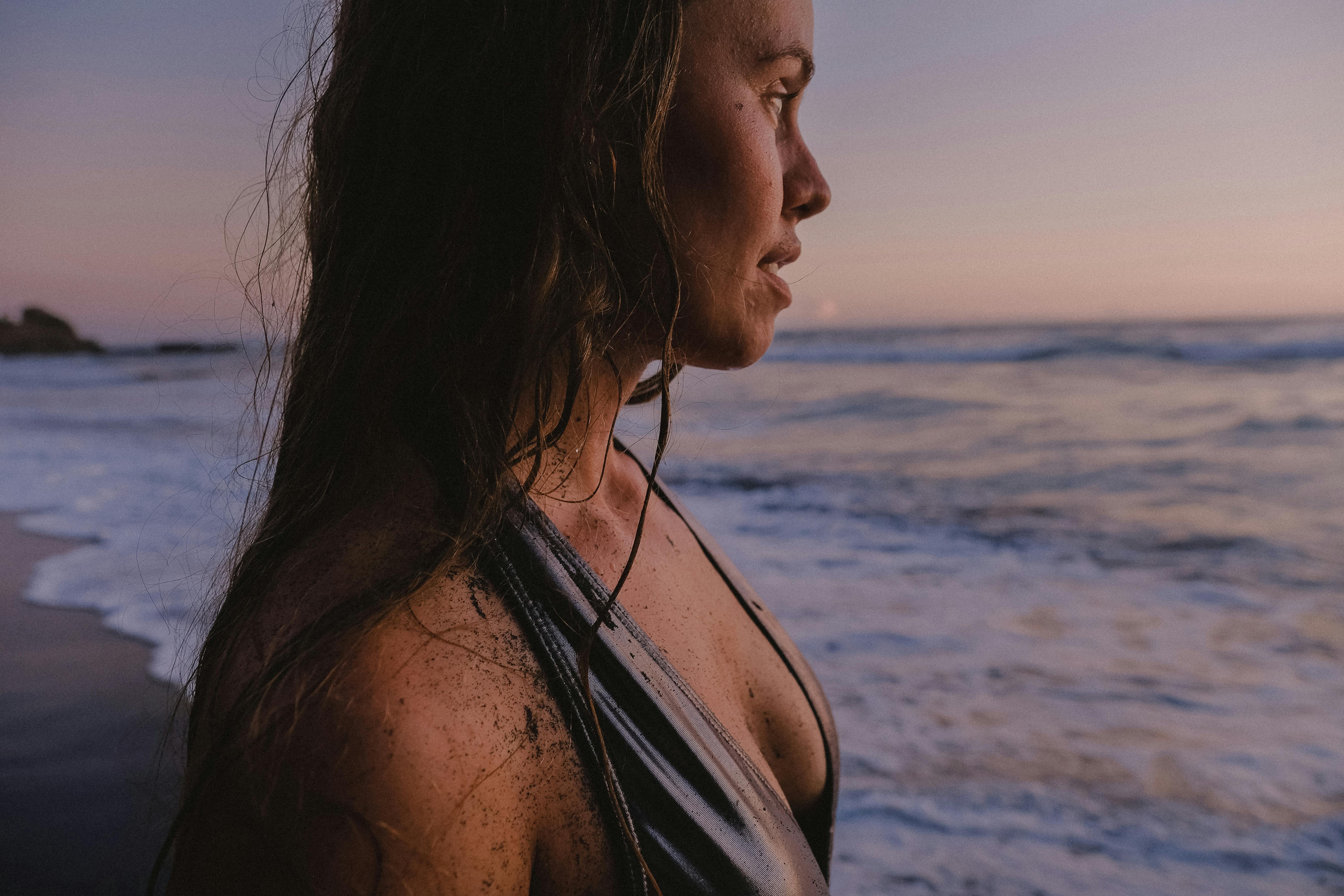 Portrait of a woman in a swimsuit standing at the beach during sunset with gentle waves in the background.