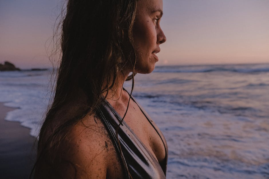 Portrait of a woman in a swimsuit standing at the beach during sunset with gentle waves in the background.