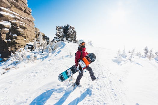 A snowboarder stands atop snowy mountains with bright blue sky and rugged cliffs.