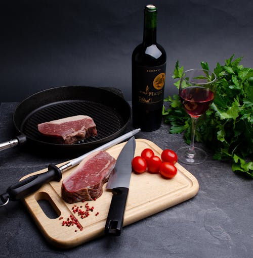 High angle of meat steaks placed on cutting board and grill pan near glass of wine parsley and tomatoes