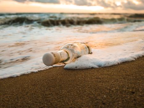 Ground level of empty glass bottle thrown on beach of foaming ocean in evening