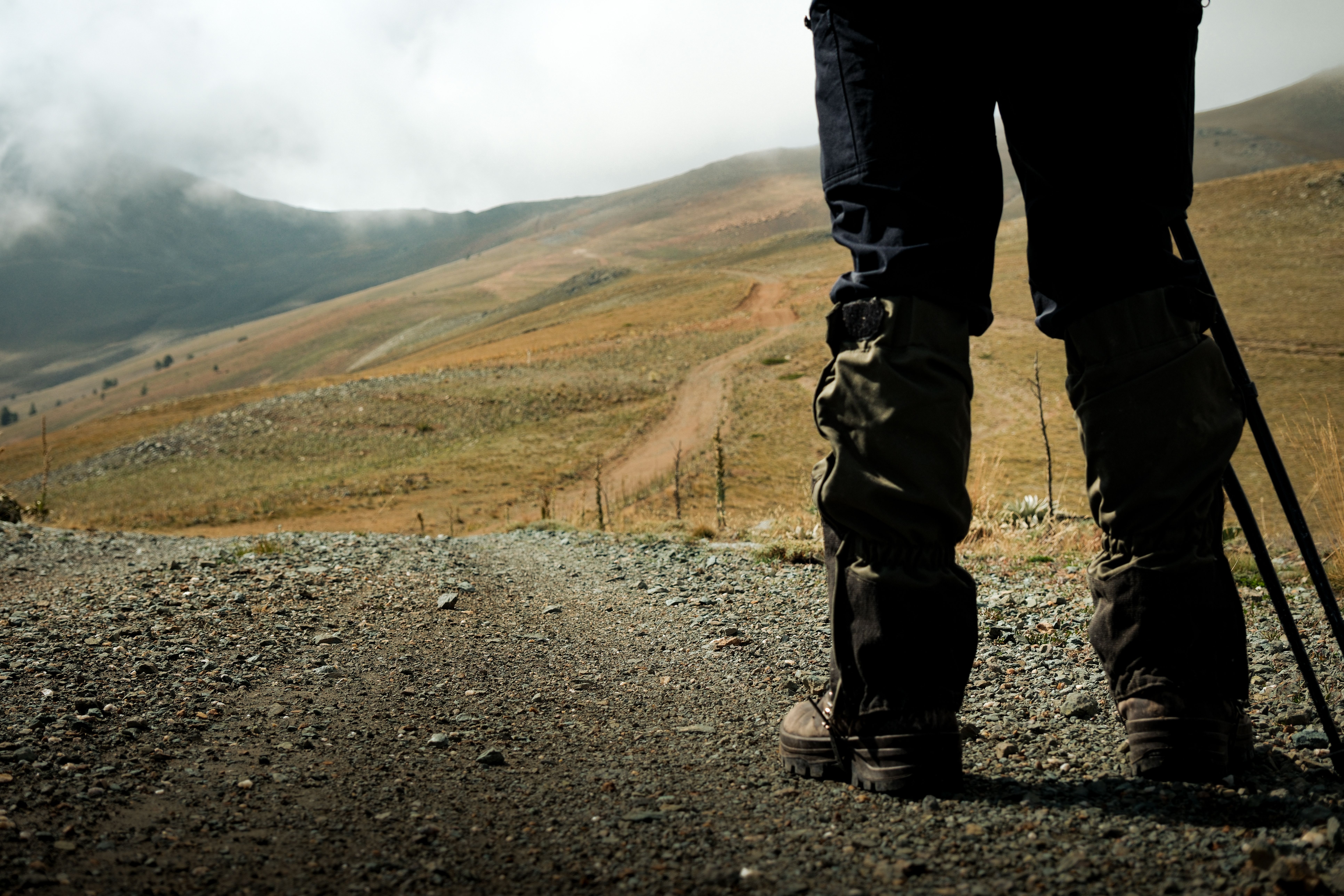 A hiker stands on a rocky mountain trail with walking poles, facing vast hilly landscapes.