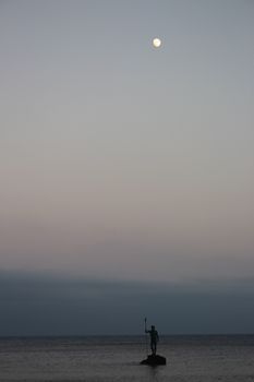 宁静的夜空中，一座孤独的渔夫雕像矗立着，头顶明月，营造出宁静的沿海景象