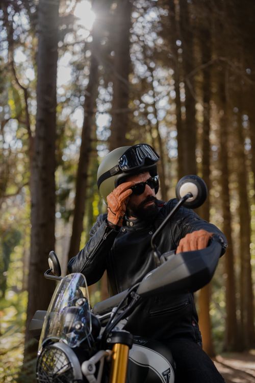 Free A motorcyclist wearing leather and a helmet adjusts sunglasses in a sunlit forest. Stock Photo