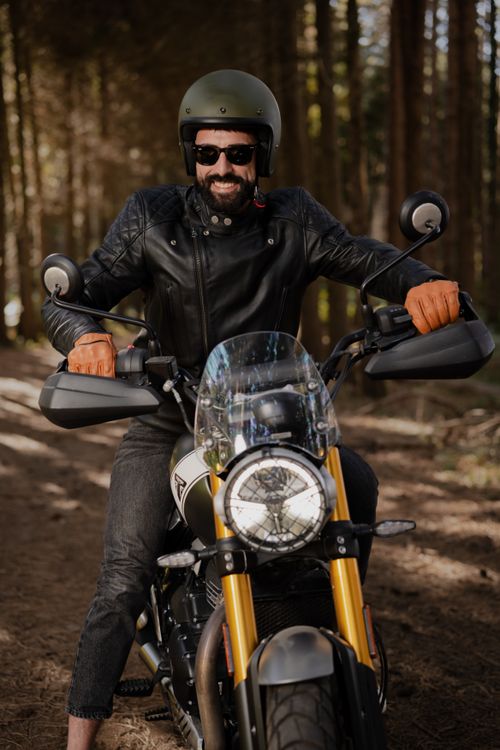 Free Smiling man in leather jacket and helmet riding a motorcycle on a forest path, showcasing adventure and freedom. Stock Photo