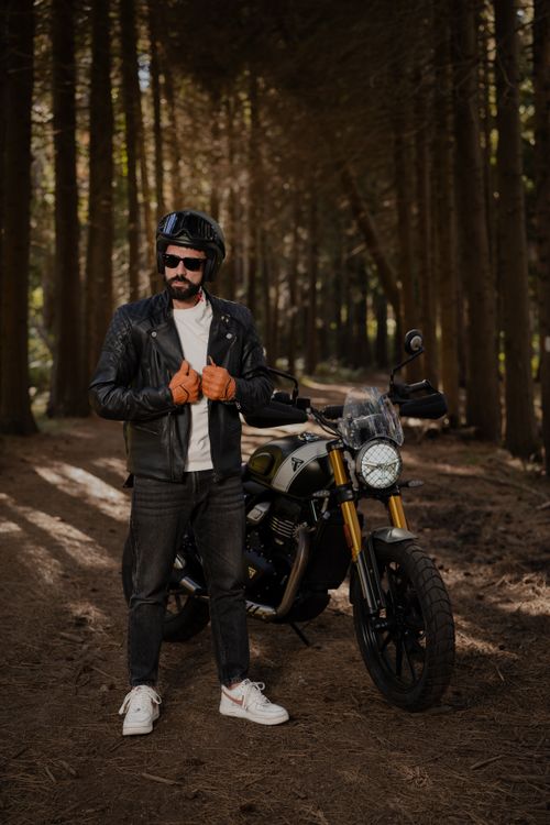 Free Man in leather jacket poses with motorcycle in a forest, showcasing an adventurous lifestyle. Stock Photo