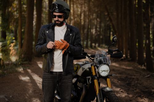 Free A motorcyclist in a leather jacket takes a break in a serene forest path during daytime, adjusting gloves. Stock Photo