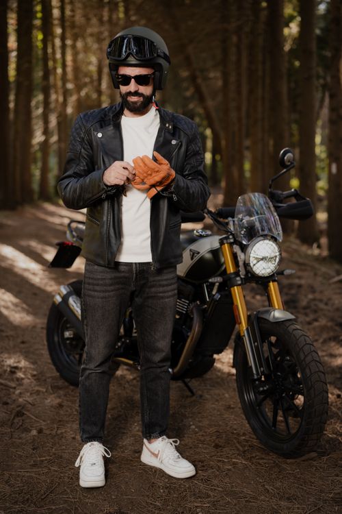 Free Motorcyclist wearing leather jacket and helmet, standing beside motorcycle in forest. Stock Photo