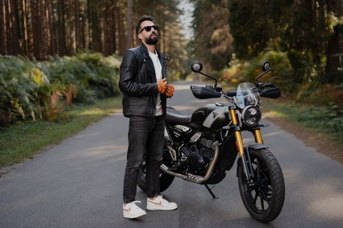 Free A stylish man in a leather jacket stands confidently beside a motorcycle on a serene forest road. Stock Photo