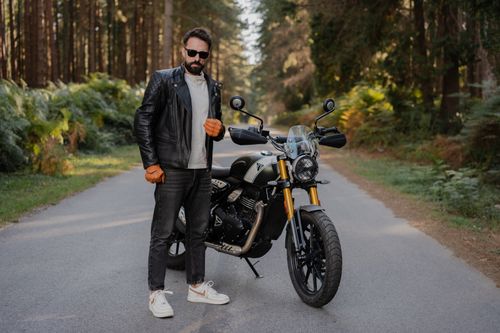 Free A man in a leather jacket stands with a motorcycle on a scenic forest road. Stock Photo