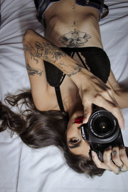 Sensual woman with tattoos lying on bed with photo camera